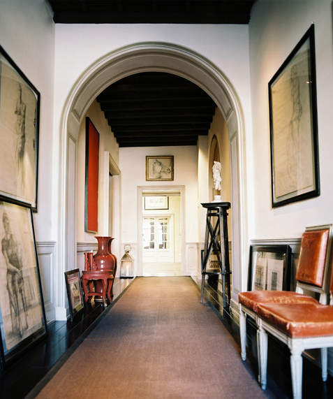 Foyer with arched entryway, dark wood floor, a long rug, two white chairs with leather seats and back and lots of sketches in black frames on the wall