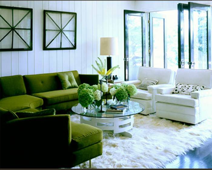 Living room with a green velvet sectional sofa, white beadboard walls, two white armchairs, a round coffee table with a white base and glass top, wood floor and a furry white rug