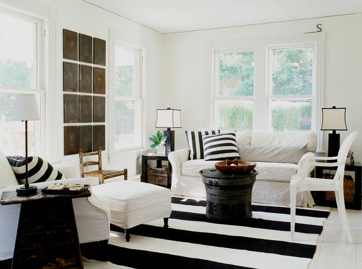 Living room in a cottage with black and white striped rug, white sofa, armchair and ottoman with black and white striped accent pillows and a black coffee table