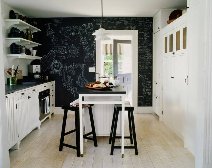 Black and white kitchen with floating shelves, white cabinets and drawers, black counter tops, an island with white legs and a black top surrounded by black stools and a floor to ceiling chalkboard