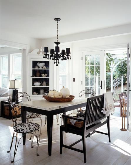 Black and white dining room in a cottage with a light wood floor, table with black legs and a marble top, black chairs with white and black graphic print seats, a dark wood bench, black chandelier, white cabinet and french doors