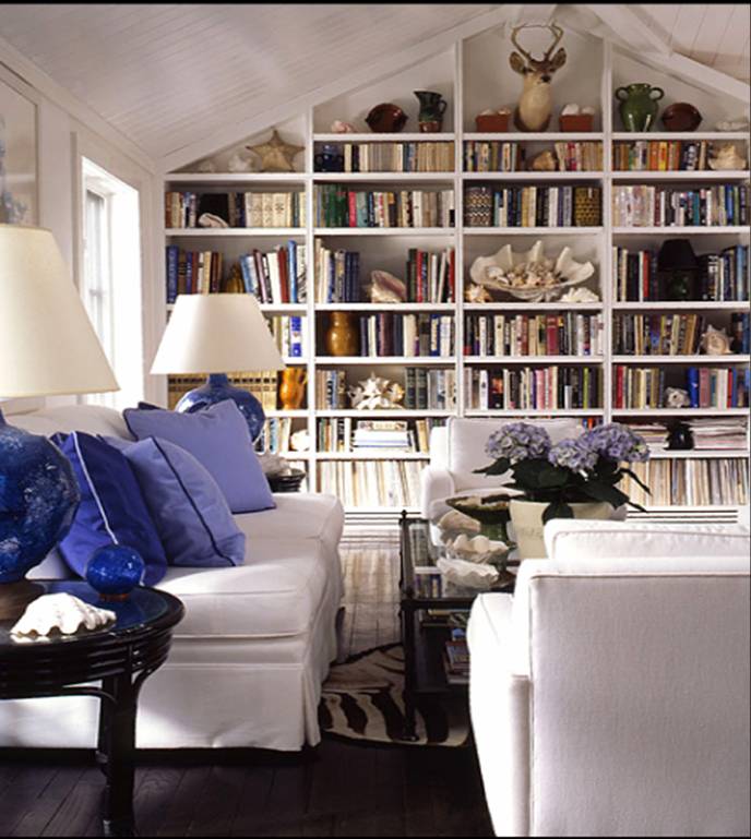 Living room with built in bookshelf full of books, dark wood floor, white sofa and matching armchairs with purple accent pillows, a zebra print rug and two lamps with blue bases and white shades on round dark wood side tables