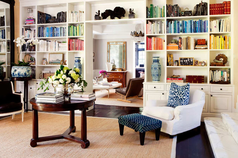 Living room with built in bookshelves full of books arranged by color, dark wood floor, white armchair with an ikat accent pillow and an upholstered blue ottoman with black legs, a sisal rug and a round wood table holding more books and a flower arrangement