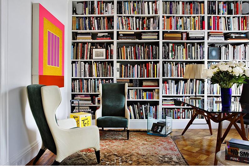 Library with floor to ceiling bookshelf full of books, parquet wood tiling, two wing back armchairs, a Moroccan rug, and a coffee table with wood legs and a glass top