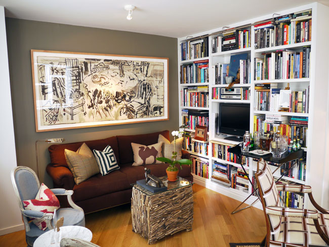Living room with floor to ceiling built in bookshelf stuffed with books and a tv, a brown sofa, a coffee table made out of twigs with a glass top, wood floor, grey walls, and a powder blue Louis XIV chair with a red and white accent pillow