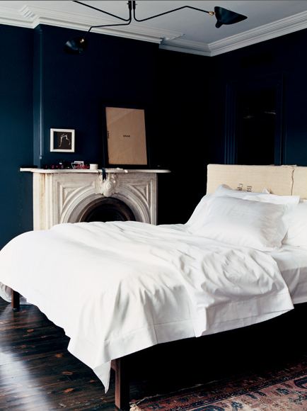 Bedroom with navy walls, a white bed, marble fireplace with a mirror on the mantel, dark wood floor, molded ceiling detail and a Turkish rug