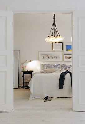 White bedroom with light wood floor, black nightstand with a metal desk lamp, blue accent pillows on the bed and a light bulb chandelier