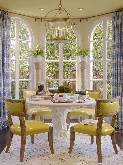 Breakfast nook with large arched windows, blue graphic print curtains, a white table with carved base, wood chairs with yellow pvc seats and backs and nail head trim