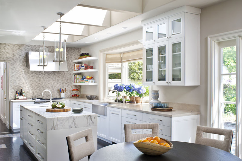 Bright kitchen with skylight, pendant lights, white cabinets and drawers with long silver pulls, marble counter tops, wood floor and a round table surrounded by upholstered chairs