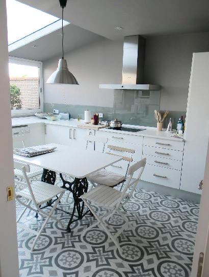 Kitchen with skylight and large picture window, stainless pendant light, white table with iron legs surrounded by by white chairs, a Moroccan tile floor and white drawers with long silver pulls