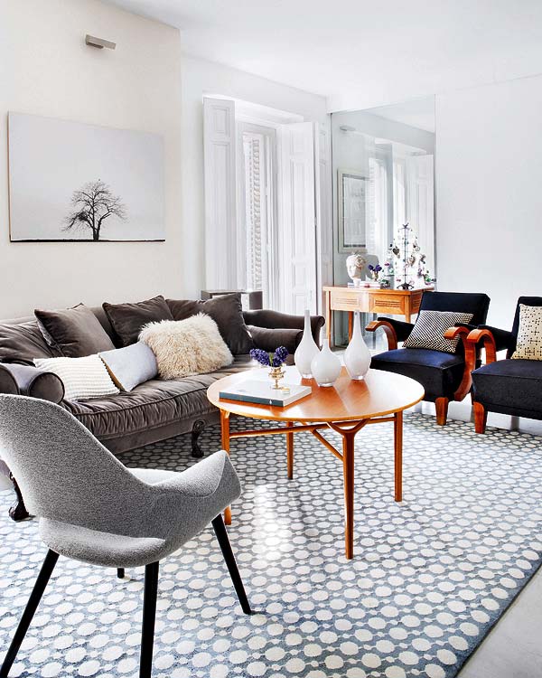 Living room in an apartment with a geometric print rug, dark blue-grey velvet sofa, two blue armchairs, a grey upholstered chair, and a round wood coffee table
