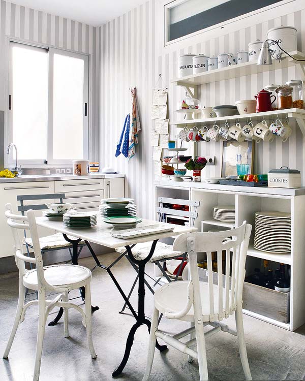 Alternative view of the apartment kitchen with grey and white striped walls, white drawers with long drawer pulls, stainless counter top, open shelving, an table with iron legs and a marble top and mismatched chairs