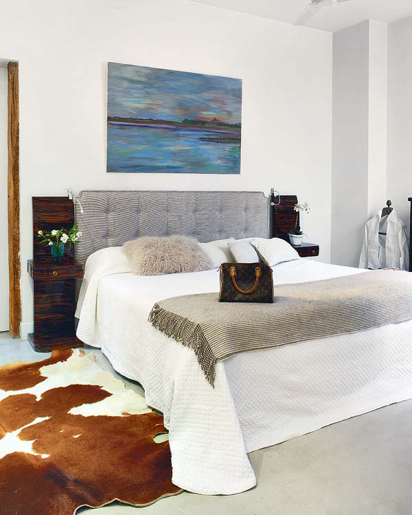 Bedroom with animal skin rug, tufted grey headboard, above the headboard is a painting of a lake, white bedding with a throw at the foot of the bed, and tall nightstands