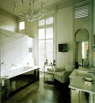 Bathroom with white chandelier, black tub, wood floor, white armchair, large windows and mirrored cabinets beneath the sink