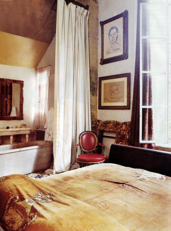 Bathroom in a loft with a stand alone marble tub, floor length curtain, red Louis XIV chair, a bed and framed sketches