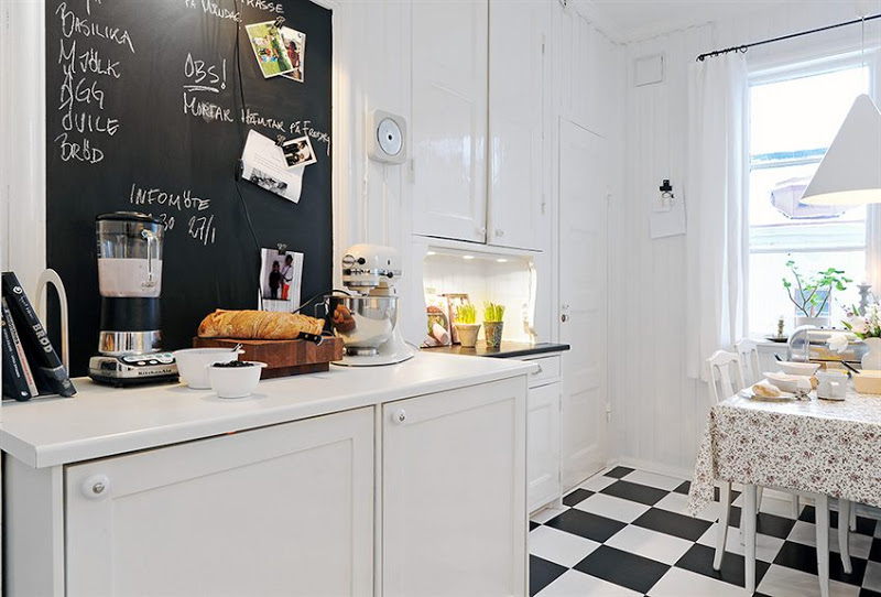 Kitchen with black and white checker floor, beadboard walls, open shelving, white pendant light over a table surrounded by white chairs and a chalkboard