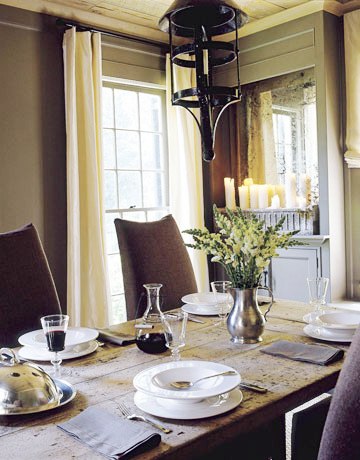 Dining room with raw wood plank ceiling, metal pendant lights, a long wood table surrounded by linen covered Parsons chairs, grey walls, and encasement windows covered by cream curtains