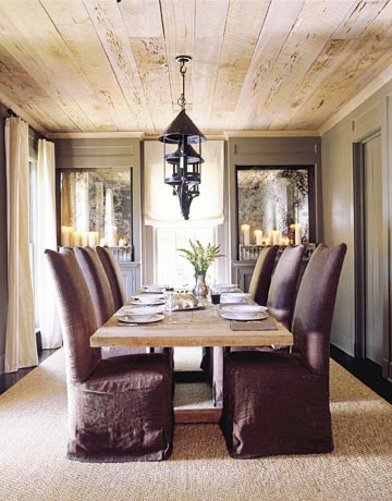 Dining room with raw wood plank ceiling, a row of metal pendant lights, a long wood table surrounded by linen covered Parsons chairs, grey walls, a dark wood floor and a sisal rug