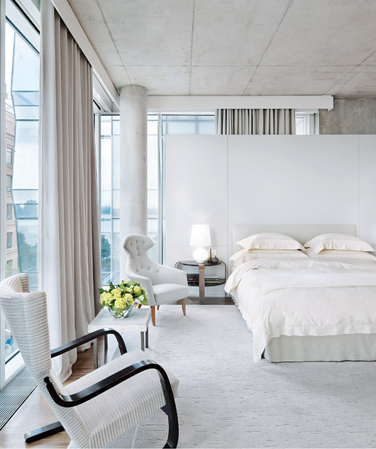 Bedroom in a NYC apartment with cement pillars, wrap around windows, a floating wall behind the bed, white upholstered headboard, a tufted armchair, a side table with a marble top, wood floor with a large area rug