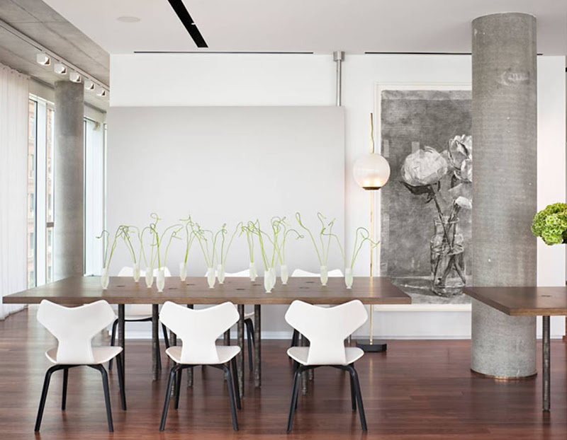 Dining room in a NYC apartment with cement pillars, wood floor, long wood table surrounded by white chairs and a large sketch of flowers in a vase on the wall