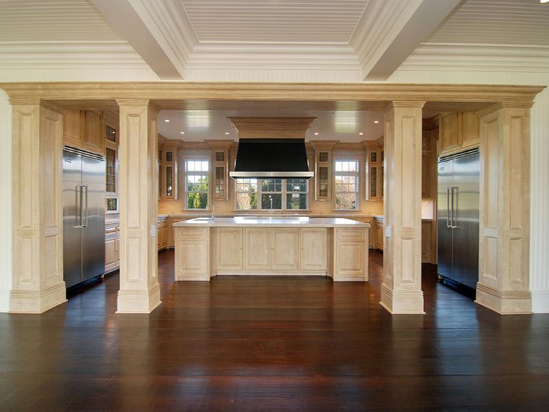 Empty kitchen in a Hampton's farm with coffered ceiling, wood floor, stainless appliances light wood cabinets, drawers and columns and a matching island
