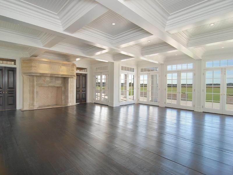 Empty living or family room in a Hamptons farm with coffered ceiling, large windows, French doors, wood floor and a massive fireplace