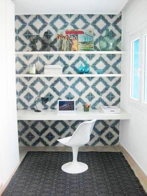 Blue and white diamond cement tiles behind a white desk with floating shelves. In front of the desk is a white Tulip Chair on a dark rug with a diamond pattern