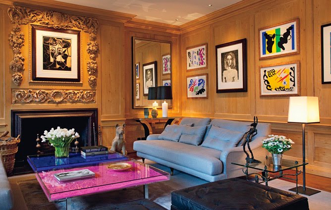 Living room with Yves Klein's Table Bleue and Table Rose, wood paneled walls covered in frames pieces of art, a fireplace with an intricately carved mantel, a leather sofa, a black tufted leather ottoman and a glass side table