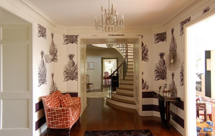 Hallway in a remodeled and restored Regency home by Kevin Oreck with striped brown and white wainscoting, an orange and white graphic print settee and white wallpaper with a botanical print of a pineapple 